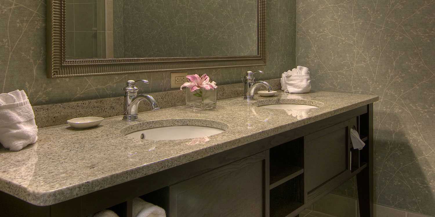 Salle de bain d'hôtel avec double vasque, comptoir en granit, deux robinets chromés, miroir, serviettes blanches roulées et un lys rose