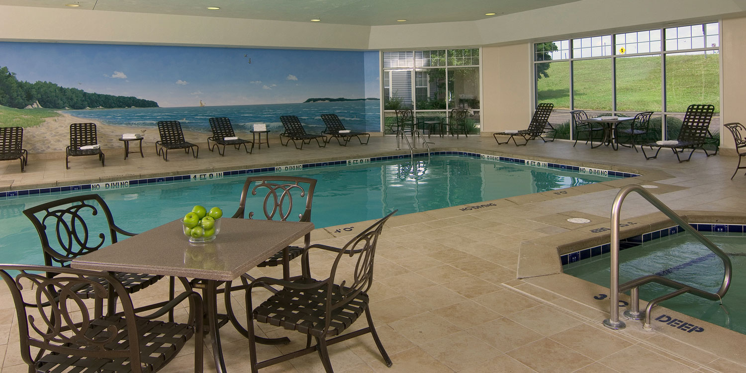 Espace piscine intérieure avec chaises longues, petites tables, bain à remous et fresque murale de plage, tons bleus et beiges